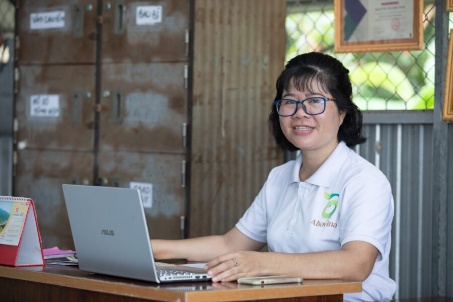 A woman of Vietnamese heritage with black bobbed hair, glasses and a white t-shirt is sat in her office working at a laptop. She is looking at the camera and smiling.