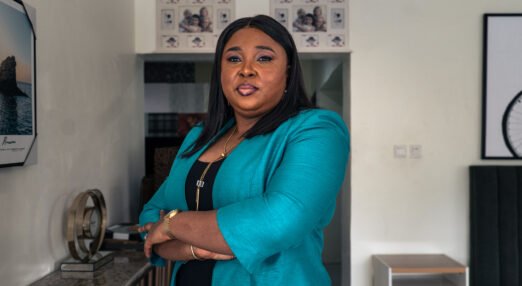 Portrait of Glory Omoregie, a Black woman with shoulder-length straight dark hair. She is wearing a blue blazer over a black top and stands in a room looking powerful with her arms folded, looking at the camera.