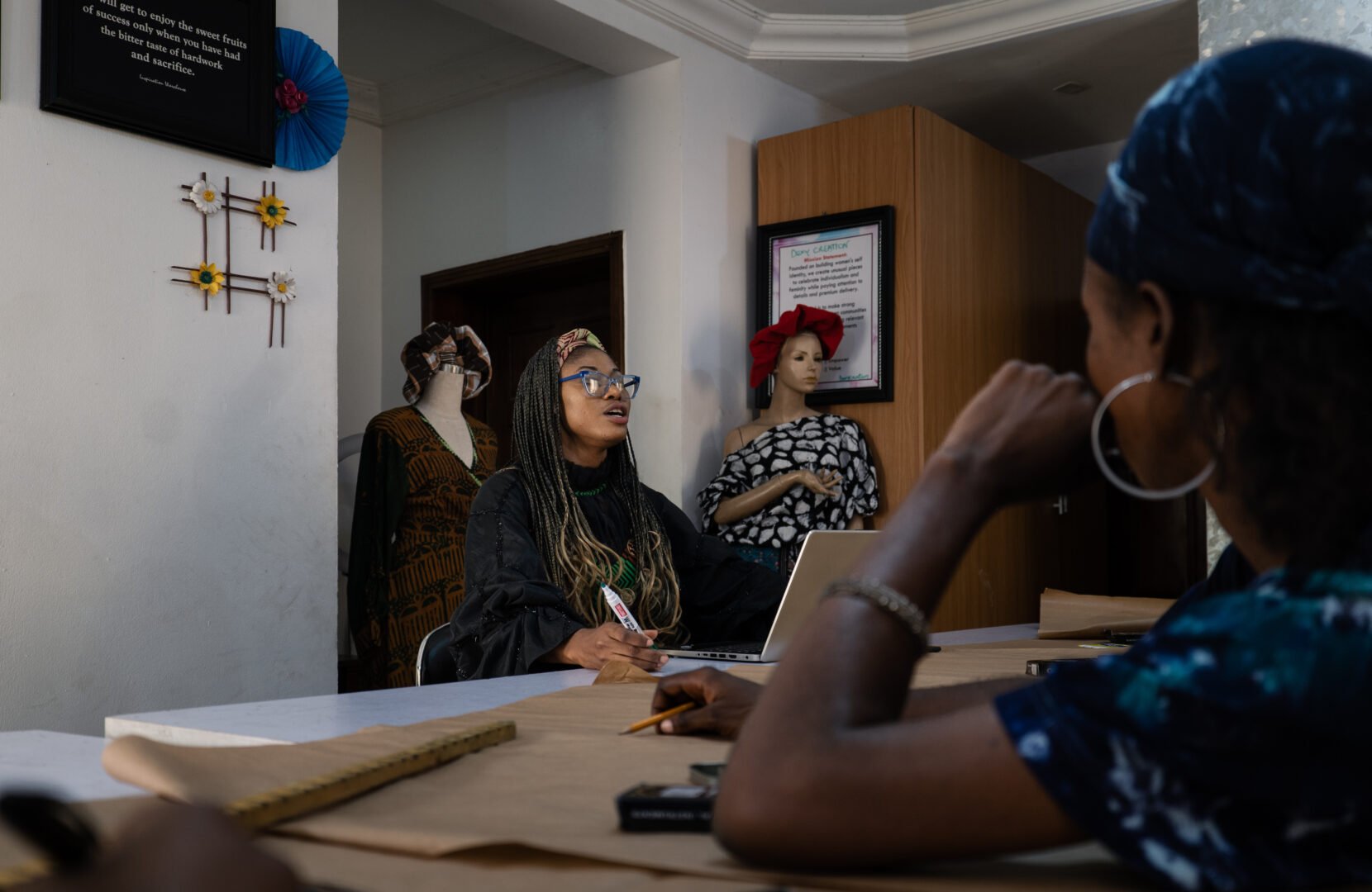 Adaeze Onu, a Nigerian woman with braids, glasses and a colourful headband, sits with her staff around a meeting table in her office. There are mannequins in garments standing behind her and she is holding a marker pen and speaking.