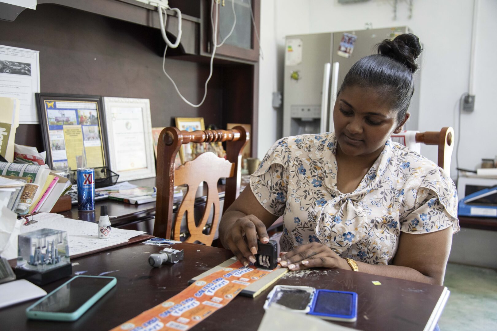 Radhika Basdeo, a Guyanese woman of south Asian heritage, sits at a desk in her office. Her hair is up in a bun and she is wearing a floral blouse. She is date-stamping a batch of labels by hand.
