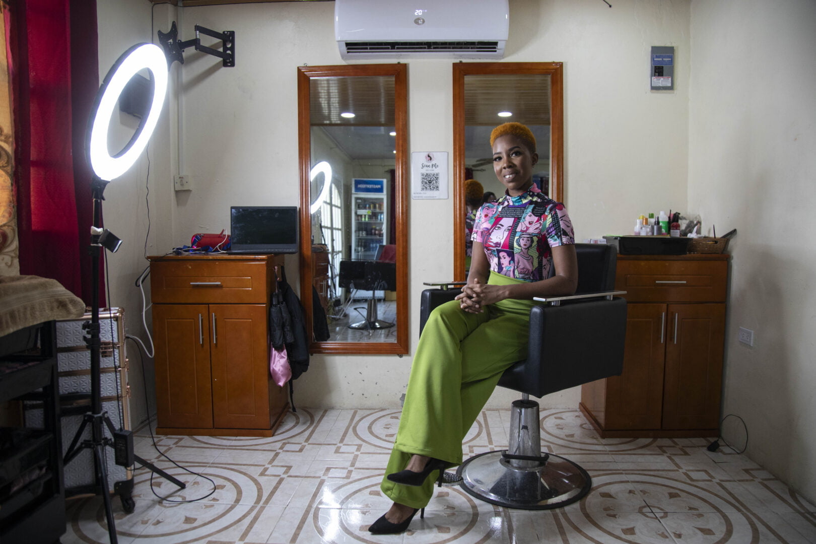 Latoya Rigby, a Black woman with very short, organe hair, poses in a brightly coloured patterned top and bright green trousers. She is sitting in her hair salon, in a hair cutting chair, with a bright ring light illuminating her, and is smiling at the camera.