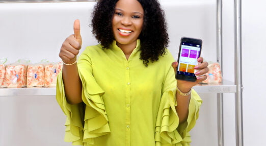 A Nigerian woman with shoulder-length curly black hair wearing a bright yellow blouse is standing in an office space. She is smiling at the camera and giving a thumbs-up whilst her other hand holds up and shows the HerVenture app on her smartphone.
