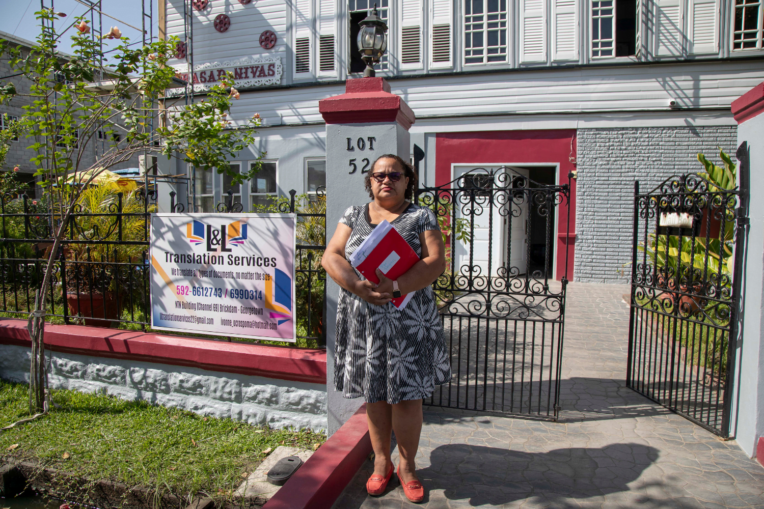 A woman entrepreneur, Ivonne Ocrospoma, stands outside on a sunny day. Behind her is a gated business. On the gate is a sign that says "L&L Translation Services". She is wearing read shoes and a black and white dress with plant patterns all over it. Her hair is tied into a ponytail and she is carrying a red folder that is full of documents.