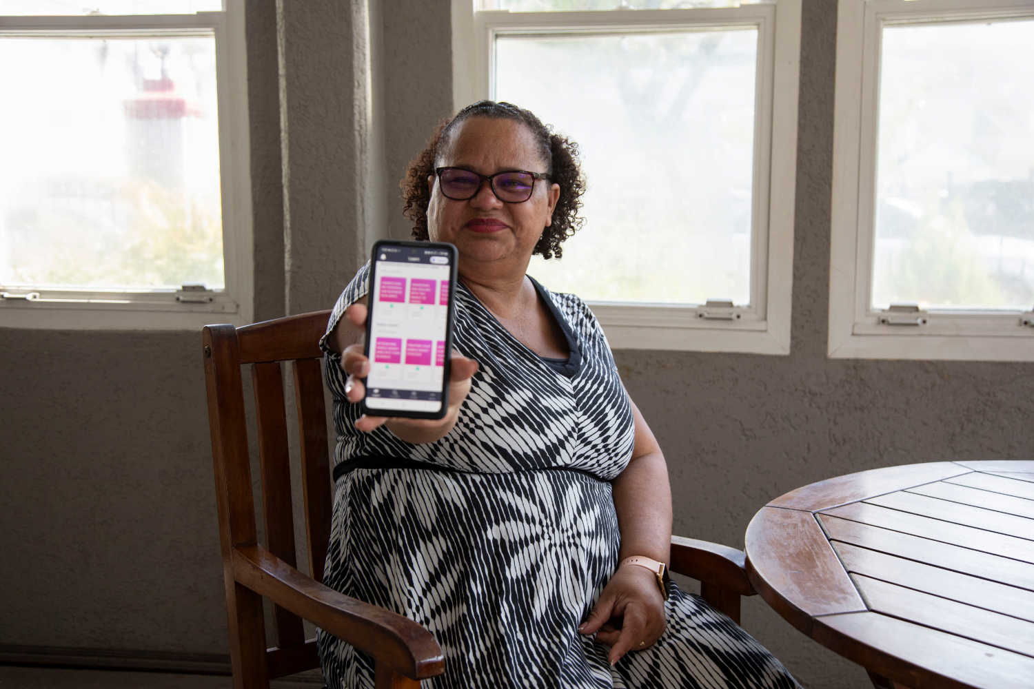A woman entrepreneur, Ivonne Ocrospoma, sits in a wooden chair in front of a window. She is wearing a black and white dress with plant patterns on it. She is holding out a phone with the HerVenture app on the screen and she is smiling.