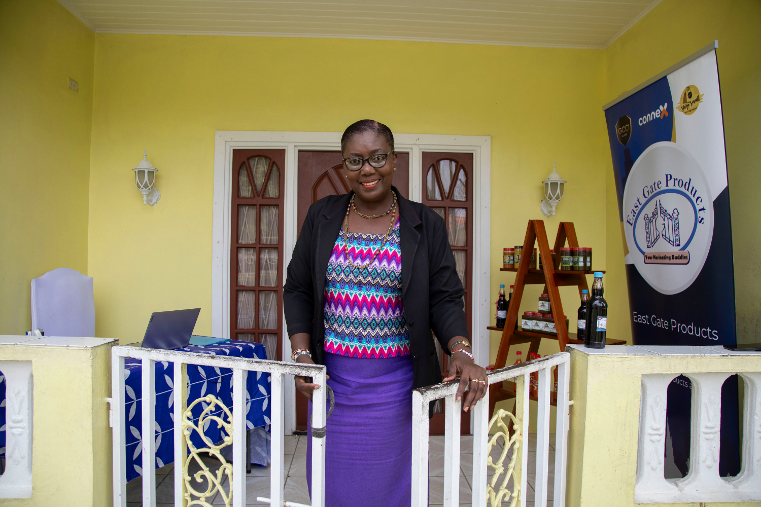 A Guyanese woman entrepreneur, Stacy Reece, stands behind a white, hip-height metal gate. Behind her is a yellow building with an open porch. On the porch is a desk with a laptop, a sign that says East Gate Products and Café and a wooden shelf with marinating sauces on it. She is wearing a top with a pink, blue and purple chevron pattern under a black blazer and a purple skirt.