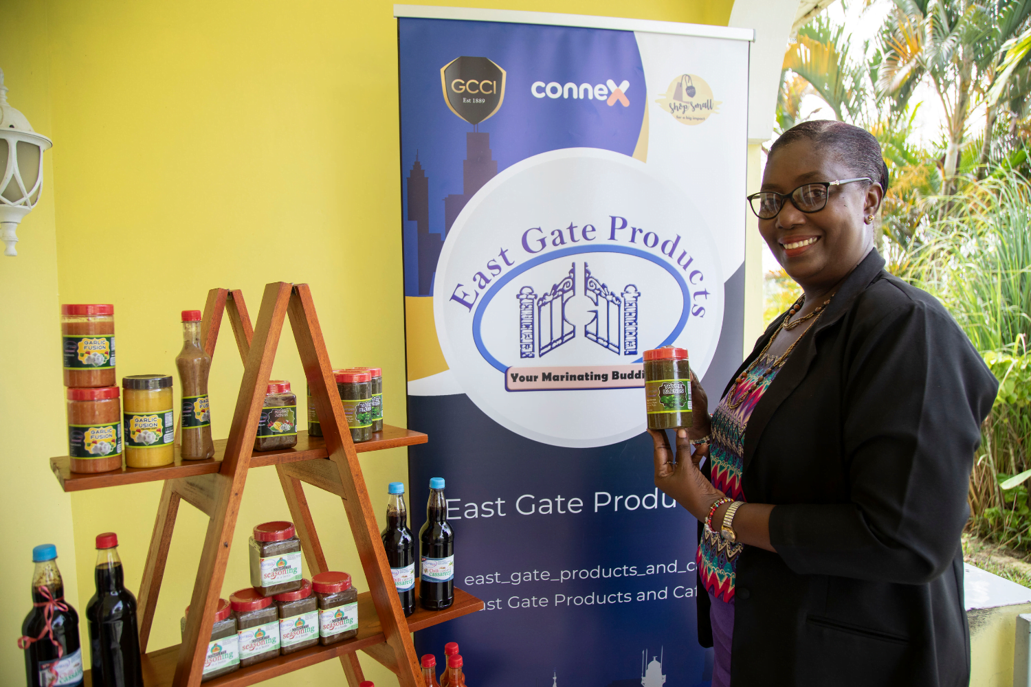 A Guyanese woman entrepreneur, Stacy Reece, stands in front of a blue and white banner sign that says East Gate Products and Café. She is smiling at the camera and holding one of her marinating sauces in a jar. Next to the banner sign is a wooden shelf with marinade sauces on it. She is wearing a top with a pink, blue and purple chevron pattern under a black blazer.