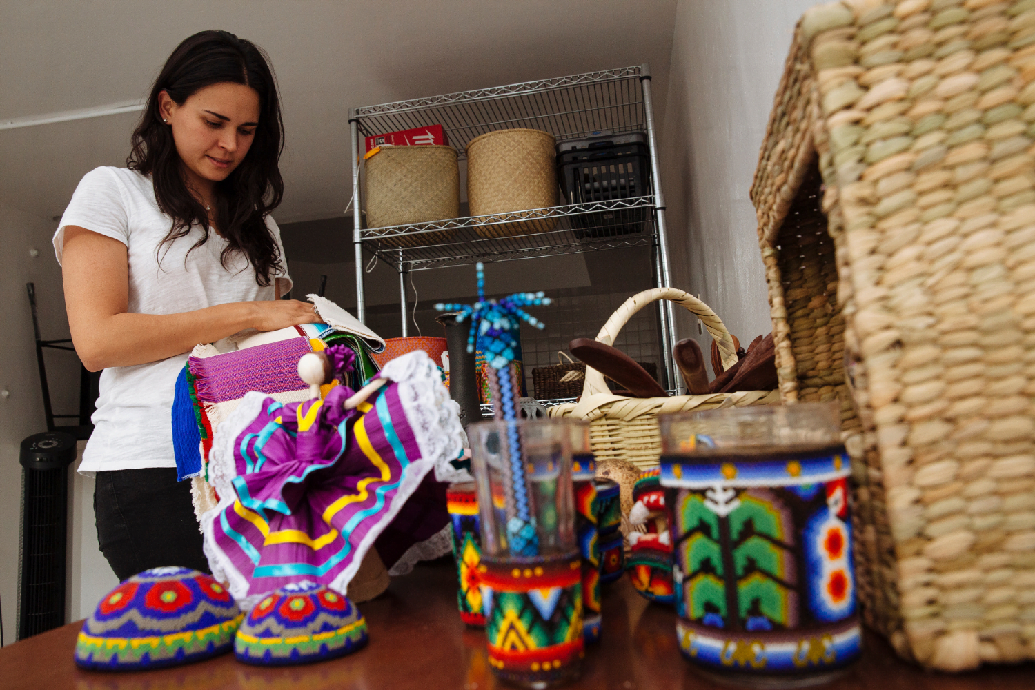 Sofia Cruz del Río Castellanos, a woman entrepreneur from Mexico works with hand beaded products. In the forefront are glass jars with intricately beaded covers. She stands in the background wearing a white t-shirt and working with a fabric that has purple, yellow and blue ribbons on it. In the background are shelves of woven baskets.
