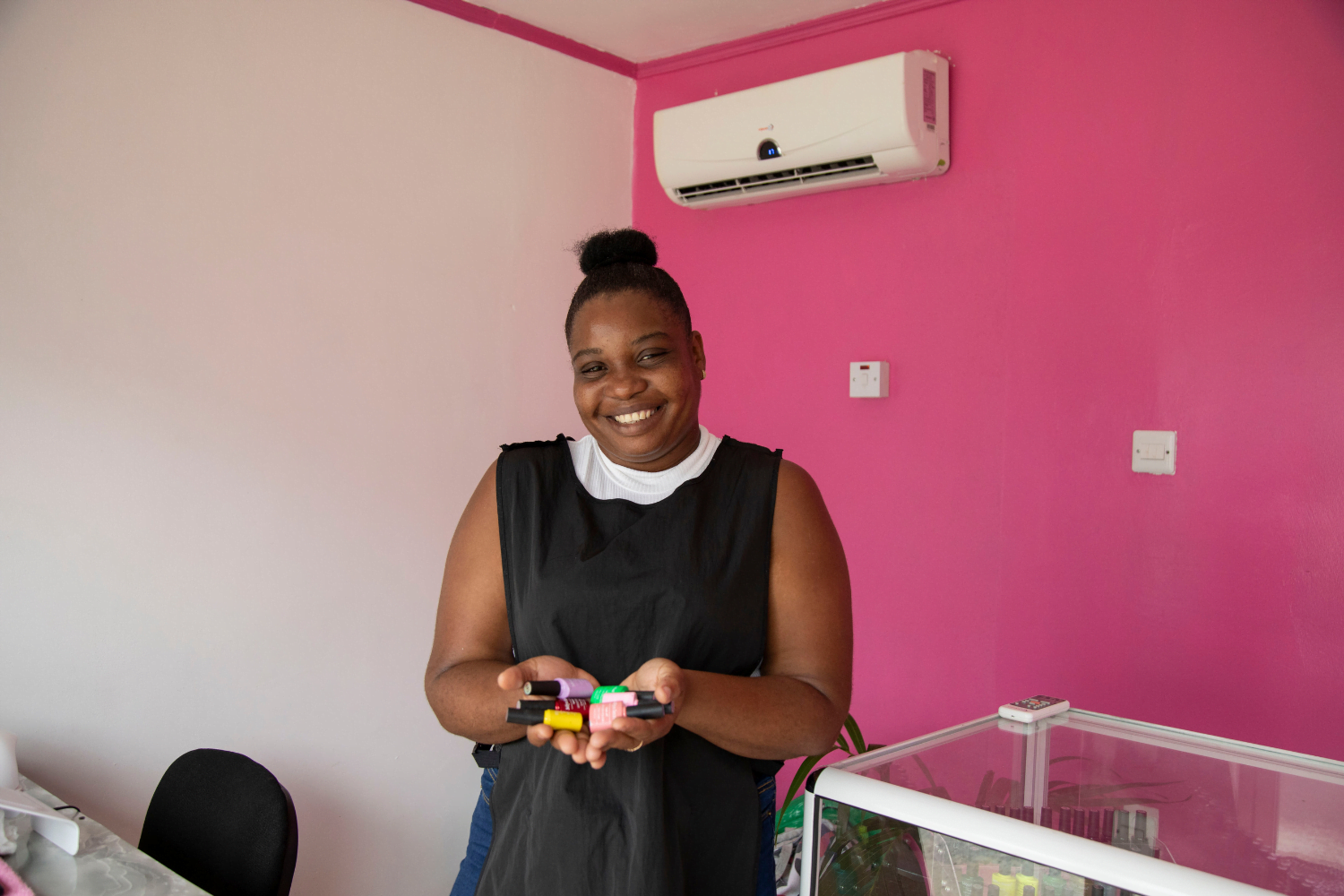 Akela Henry, a woman entrepreneur from Guyana, stands in a black smock with a white sleeveless shirt underneath in a beauty shop. She is holding many colourful nail polish bottles. Her hair is in a bun and she is smiling.