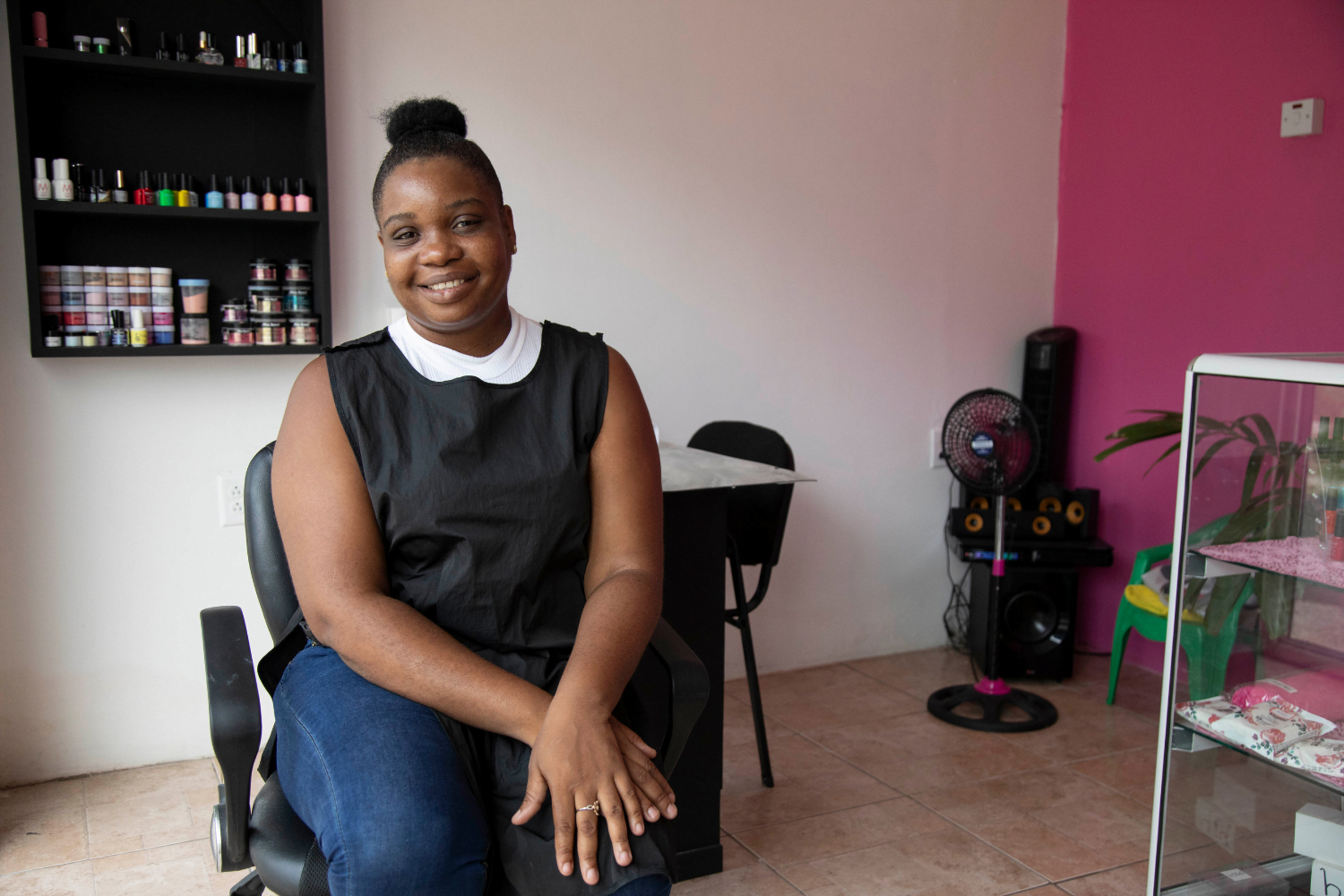 Akela Henry, a woman entrepreneur from Guyana, sits in a black smock with a white sleeveless shirt underneath in a beauty shop. She is in front of a nail station and there is a glass case nearby with many colourful nail polishes in it. In the background there is a white wall with more nail polishes on display and a pink wall. Her hair is in a bun and she is smiling.