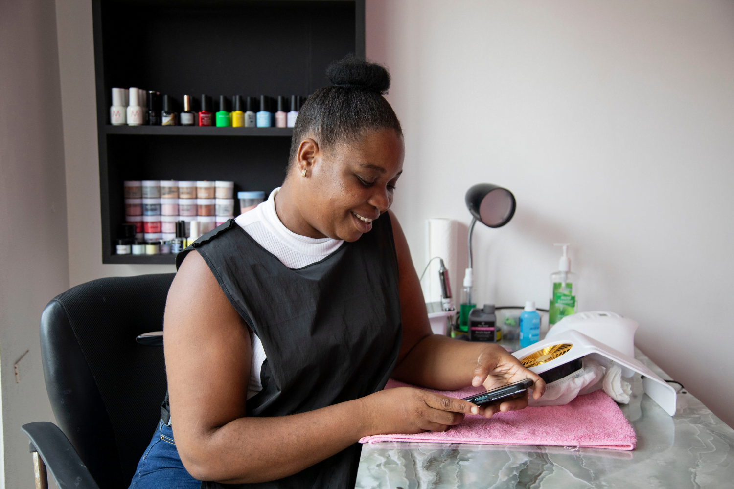 Akela Henry, a woman entrepreneur from Guyana, sits in a black smock with a white sleeveless shirt underneath in a beauty shop. She is using her smartphone. Her hair is in a bun and she is smiling.