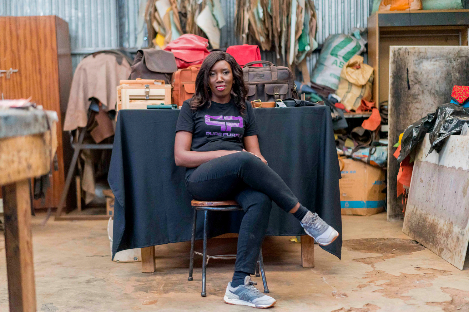A Kenyan woman entrepreneur, Yolanda Odida, sits in front of a black table with several leather bags, produced by her company Pure Purple.