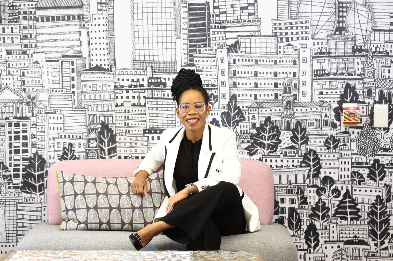 Queen Mokulubete, a Black South African woman with black hair in long braids up in a bun and glasses, She is wearing a white blazer, black blouse and black trousers. She is smiling as she poses on a pink sofa in her office in front of a wall covered in black and white line drawings.