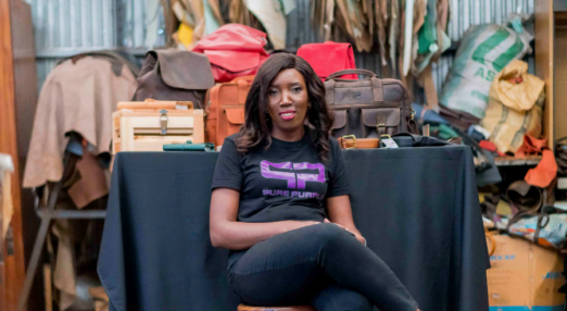 A Kenyan woman entrepreneur is sat in front of a black table with leather bags on top of it. She is wearing a T-Shirt with her company, Pure Purple's logo on it and using her phone.