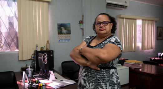 Ivonne Ocrospoma, a woman entrepreneur from Guyana stands in front of a desk with a computer and some documents on top of it. She has her arms crossed in front of her body and she's wearing a black and white dress with palm patterns all over it. She is wearing a black pair of glasses and has her hair pulled back into a ponytail.