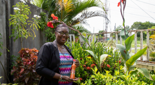 A Guyanese woman entrepreneur, Stacy Reece, poses with a jar of marinating sauce in a lush garden. Behind her are palm trees, green leafy plants, and red flowers. She is wearing a top with a pink, blue and purple chevron pattern under a black blazer and a purple skirt.