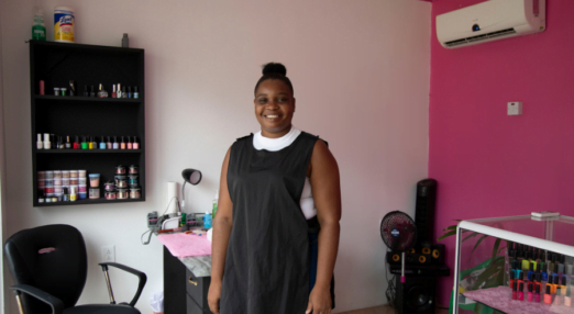 Akela Henry, a woman entrepreneur from Guyana, stands in a black smock with a white sleeveless shirt underneath in a beauty shop. She is in front of a nail station and there is a glass case nearby with many colourful nail polishes in it. In the background there is a white wall with more nail polishes on display and a pink wall. Her hair is in a bun and she is smiling.