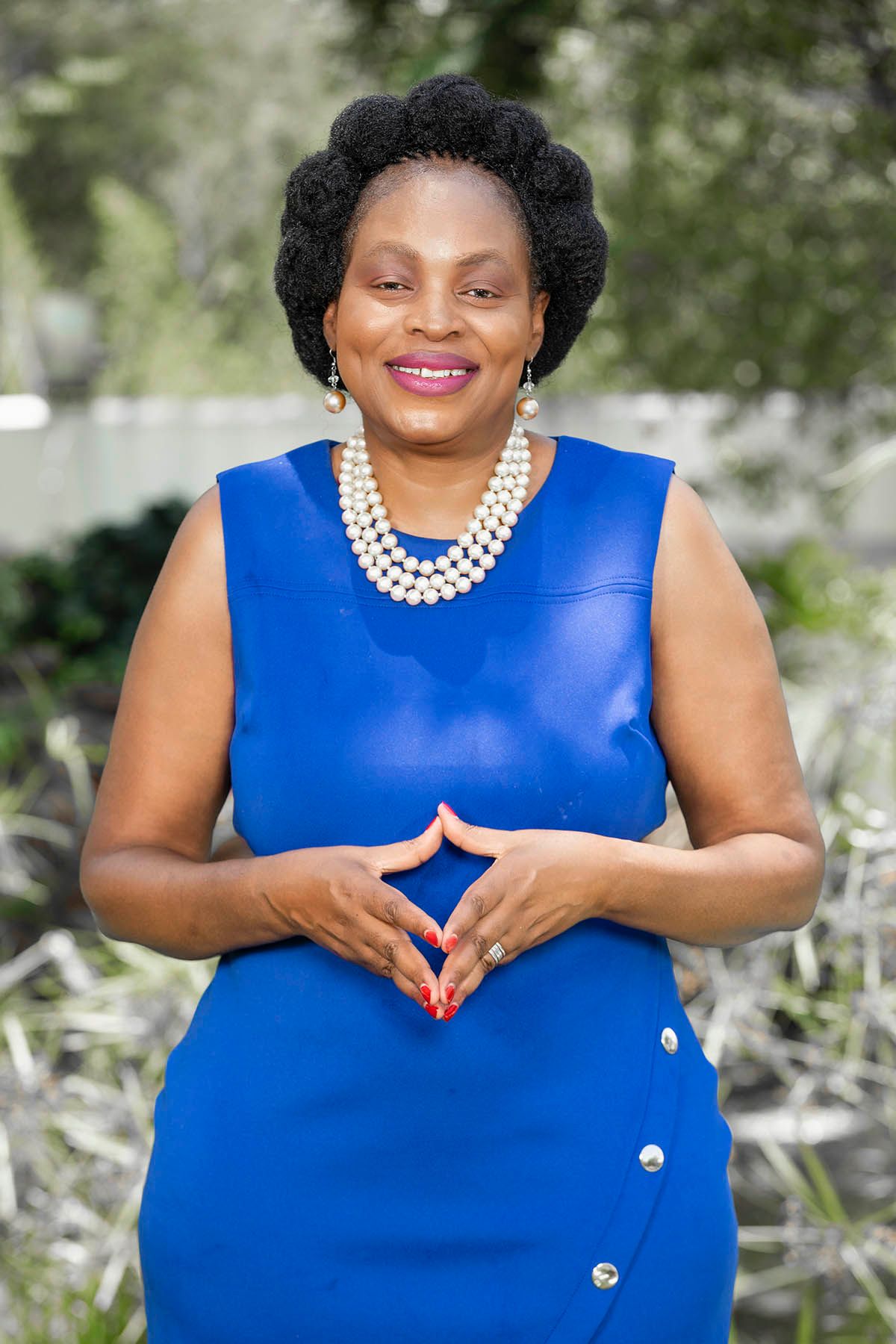 Francesca Nxedhlana, a black lady with short, natural black hair in a blue dress with pearls, smiling at the camera.