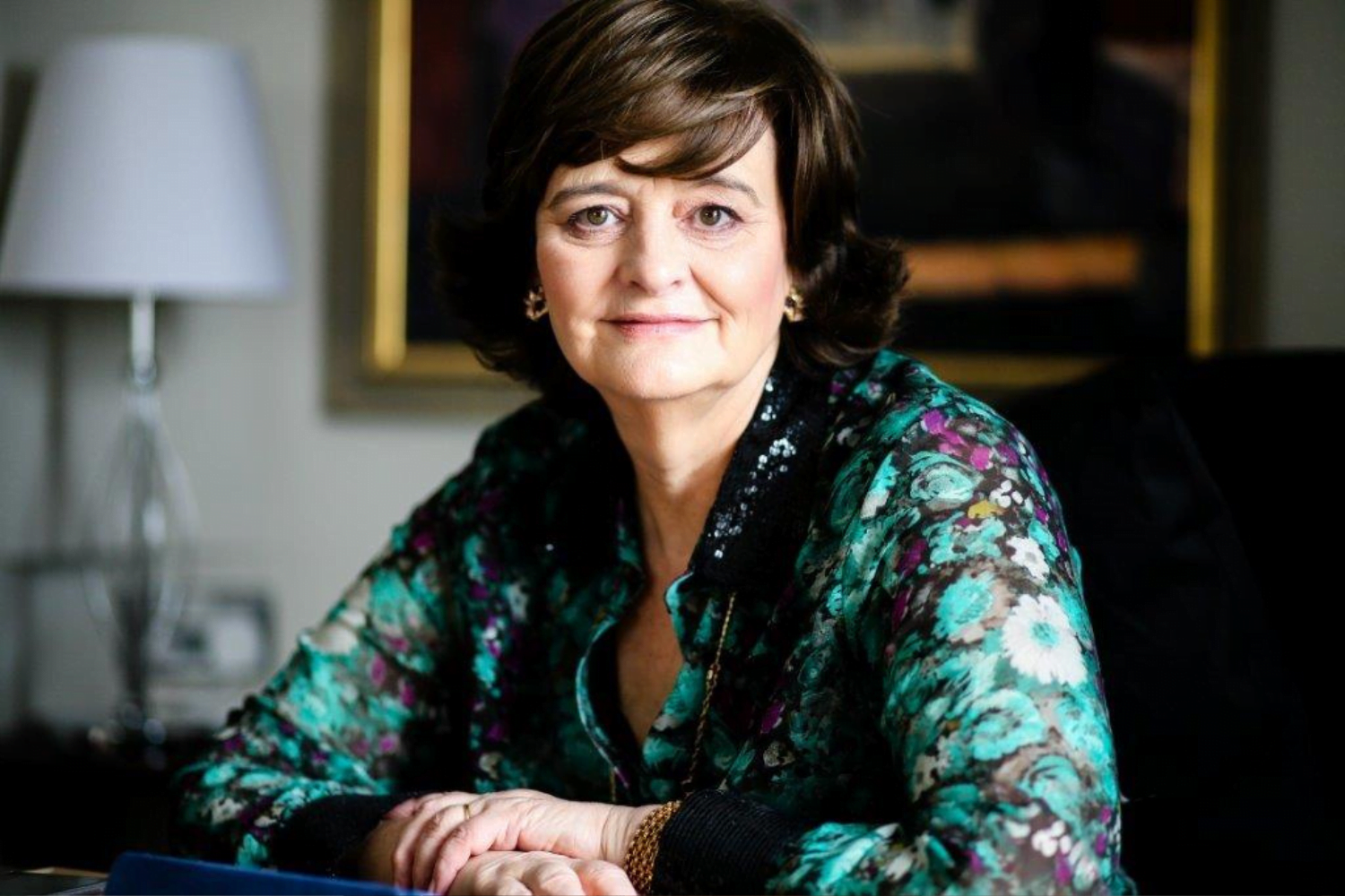 Cherie Blair, CBE KC, sits with her arms crossed on a desk. She is wearing a black blouse with teal flowers on it. In the background is a painting in a large gold frame and a white lamp.