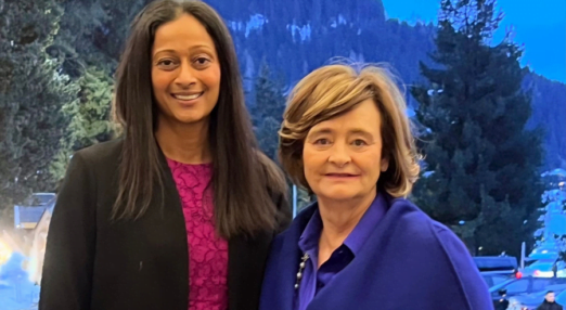 Dhivya O'Connor and Cherie Blair stand in front of a snowy mountain covered in evergreen trees. Dhivya is in a pink dress with a black blazer and Cherie is in a blue coat over a blue blouse. They are smiling.