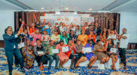 A cohort of Road to growth graduates pose for a group photo. The front row of women are sat on a bench with two rows of women behind them standing. On either end of the group, there are male training facilitators. Several people have red confetti cannons that are shooting out colourful confetti.