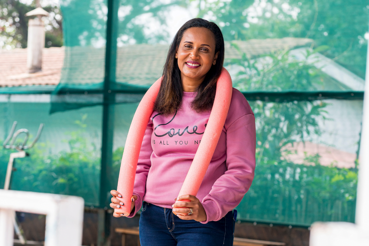 Batty Matharu, a woman entrepreneur from Kenya, stands with a pink pool noodle draped over her shoulders. She is wearing a pink sweatshirt that says "Love" and jeans. Her hair is down and she is smiling. In the background is a pool bench and a green netted wall.