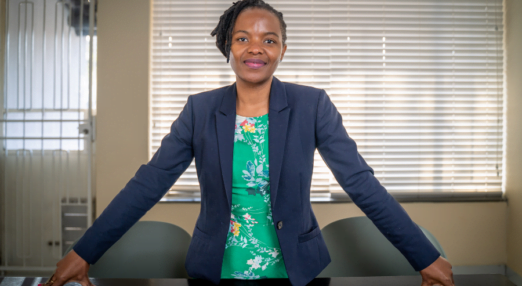Dudu Makhari, a woman entrepreneur from South Africa, poses confidently in her office. She is standing behind a wooden desk with her hands spread far apart as she leans against it. She is wearing a blue blazer and a green floral shirt. Her hair is tied up and she is smiling.