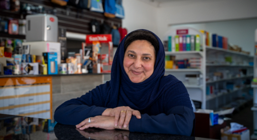 Shereen Cassim Hassim, a female entrepreneur, poses for a photograph from behind the front desk of her business. The desk is black and reflective. She is wearing a navy blue dress and hijab and smiling. In the background are office supplies on a display shelf.