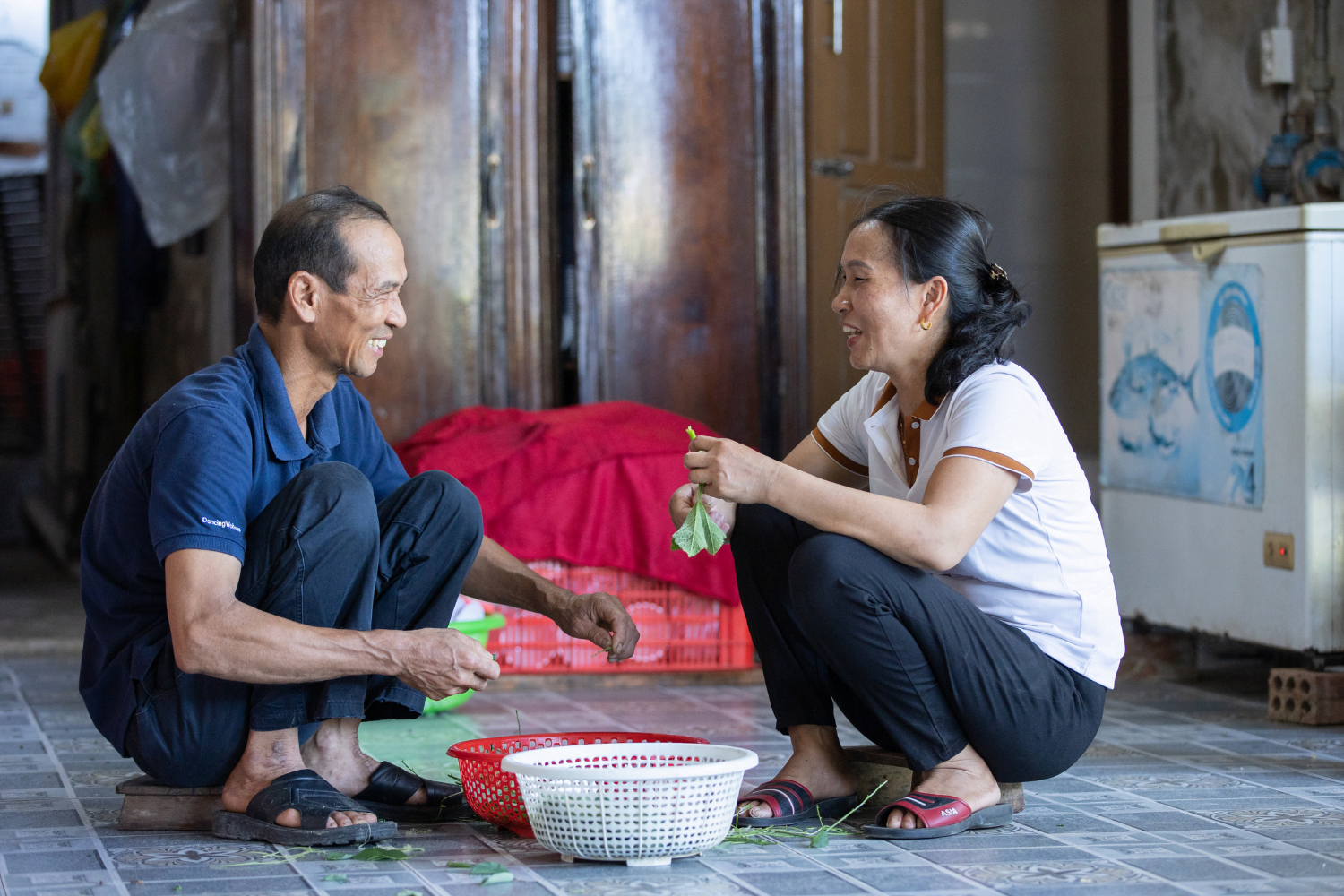 Vietnamese rice farmer Nguyen Thi Tham and her husband Nguyen Quang Trung sit on the floor together, both preparing the produce which sits in two bowls between them. They are smiling and talking to each other.