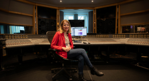 Olga FitzRoy, a UK sound engineer, sits in an office chair with her legs crossed. She is wearing jeans and black boots with a red blazer. Behind her there is sound engineering equipment.