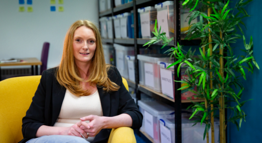 Sam Tebb, a woman entrepreneur from the UK, sits in a yellow armchair. She is wearing jeans and a white shirt with a black blazer over top. Next to her is a small potted tree and behind her are containers with informative documents for parents of disabled children.
