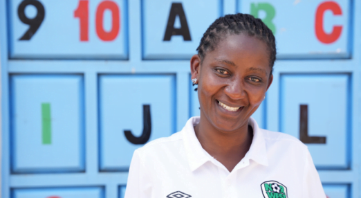 Mary Giroga, a woman entrepreneur in Kenya smiles in front of a wall with number and letter blocks painted onto it. She is wearing a white polo shirt.