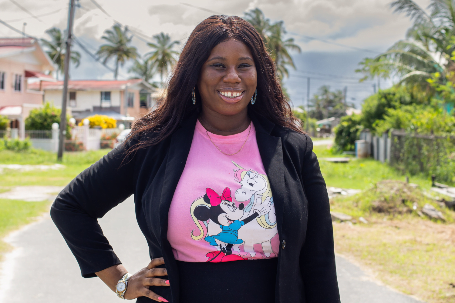 Latifah Browne, Owner and CEO of The Thrift Den, smiles confidently on a street in Georgetown Guyana. She has her hand on her hip and is wearing a blazer with a graphic tee. In the background are houses and palm trees.