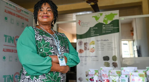 Ellah Omezi, a Nigerian woman entrepreneur, stands smiling with her arms folded in front of a table and display stand featuring her products. She is wearing a green dress.