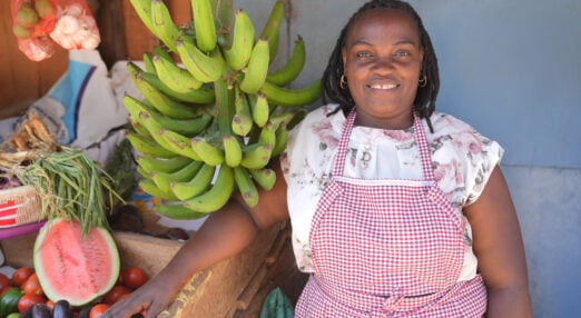 Jacqueline Muthoni Njogu at work on her fruit stand in Nairobi. She is a Black lady with medium length braided hair and is wearing a red gingham apron over a white floral dress. She is smiling at the camera with a range of fruits next to her.