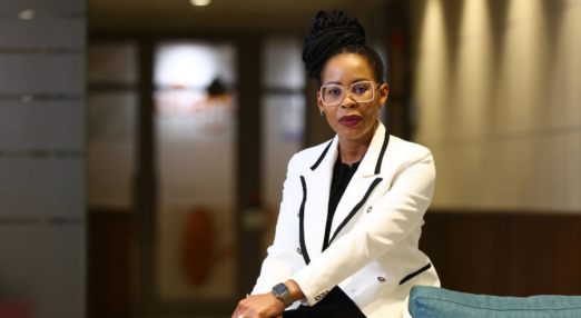 A Black woman entrepreneur sits in the foreground. She is looking seriously directly at the camera. Her background is an office. She is wearing fashionable large-framed glasses, has purple lipstick on, and is wearing a white double-breasted blazer trimmed with black.