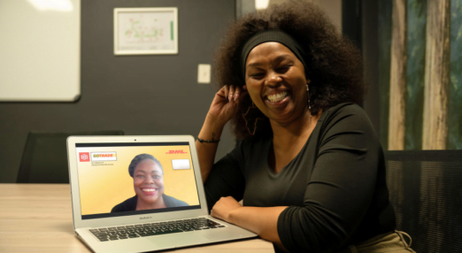 Nkeletseng Tsetsane, a woman entrepreneur in South Africa, smiles as she leans her arms against a wooden table. In front of her on the wooden table is a laptop with a woman on the screen. This is her mentor, Glory Mutai from Kenya. The two of them are facing the camera and smiling.