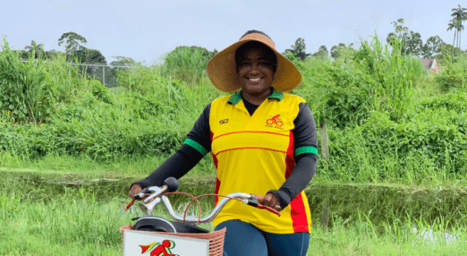 Cherry-Ann Greene, a woman entrepreneur from Guyana, is smiling at the camera and holding a bicycle by the handlebars. Her bike has a basket with 'a Ride Along logo' printed on it. The same logo is printed on her shirt.