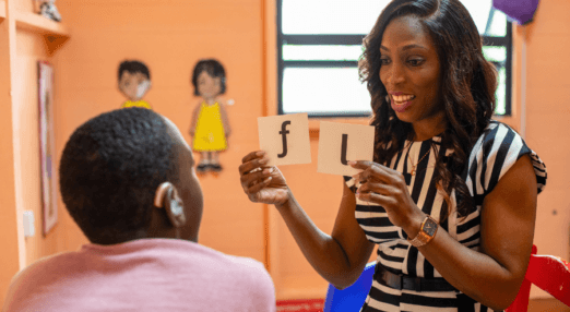 Candace Wickham shows cards with the letters 'f' and 'l' to a child in front of her, who has their back to the camera. She is looking at the child and teaching them how to say the letters. The child has a hearing aid.