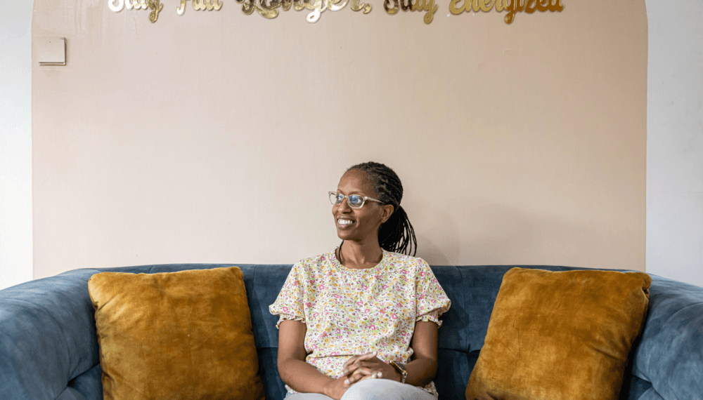 Naomi Kimuyu, a woman entrepreneur from Kenya wearing a floral top and glasses, is sat on a comfy blue sofa with gold cushions. Behind her on the wall, large gold letters spell out 'Stay Full Longer, Stay Energized'.
