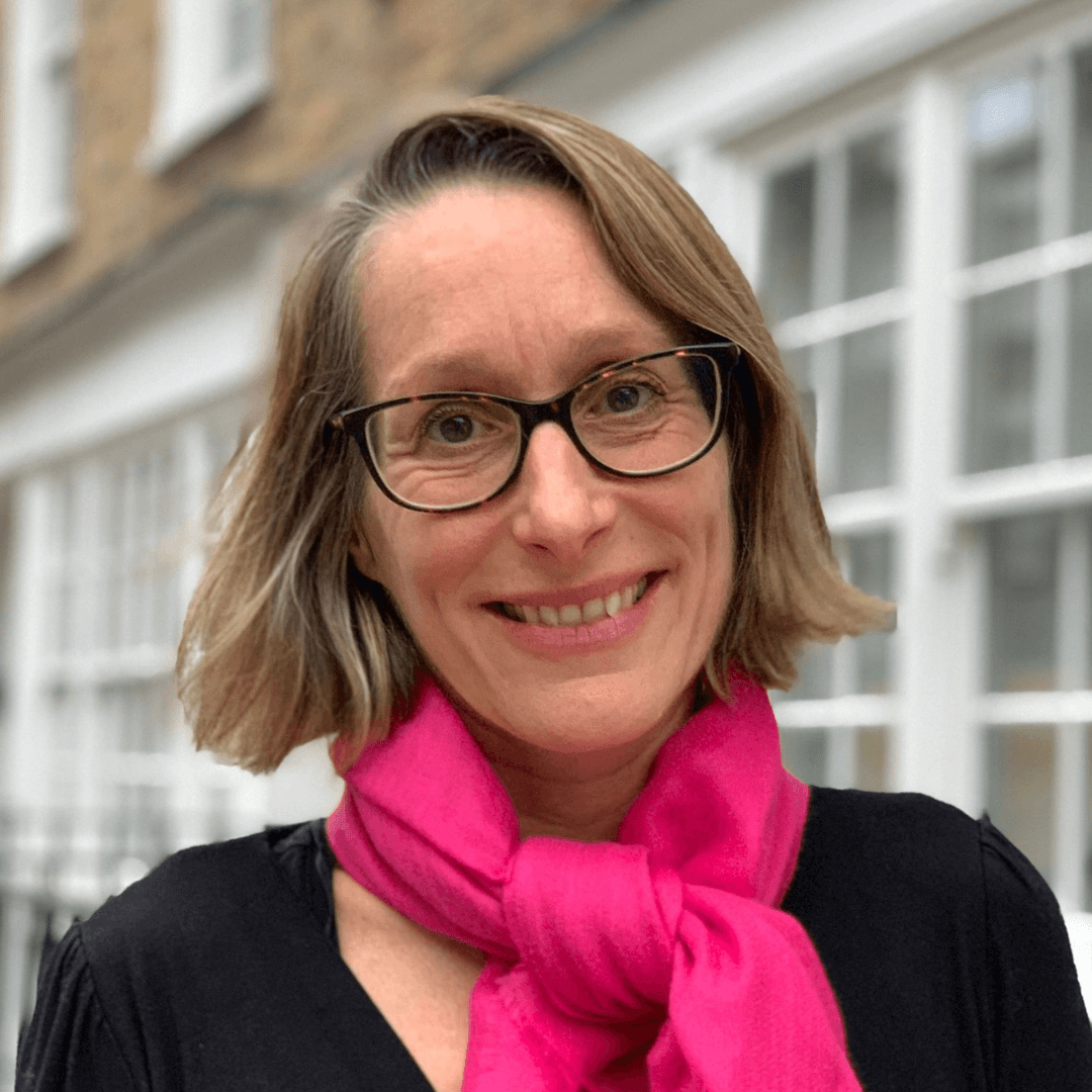 Clare Twelvetrees smiles in front of windows with white trimmings. She is wearing a black shirt, pink scarf, and black rimmed glasses. Her hair is jaw-length and blonde.