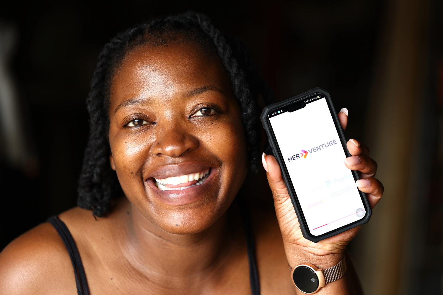 Zandile Manyisa, a woman entrepreneur in South Africa, smiles at the camera. She is holding her phone in her hand, which displays the 'HerVenture' logo.