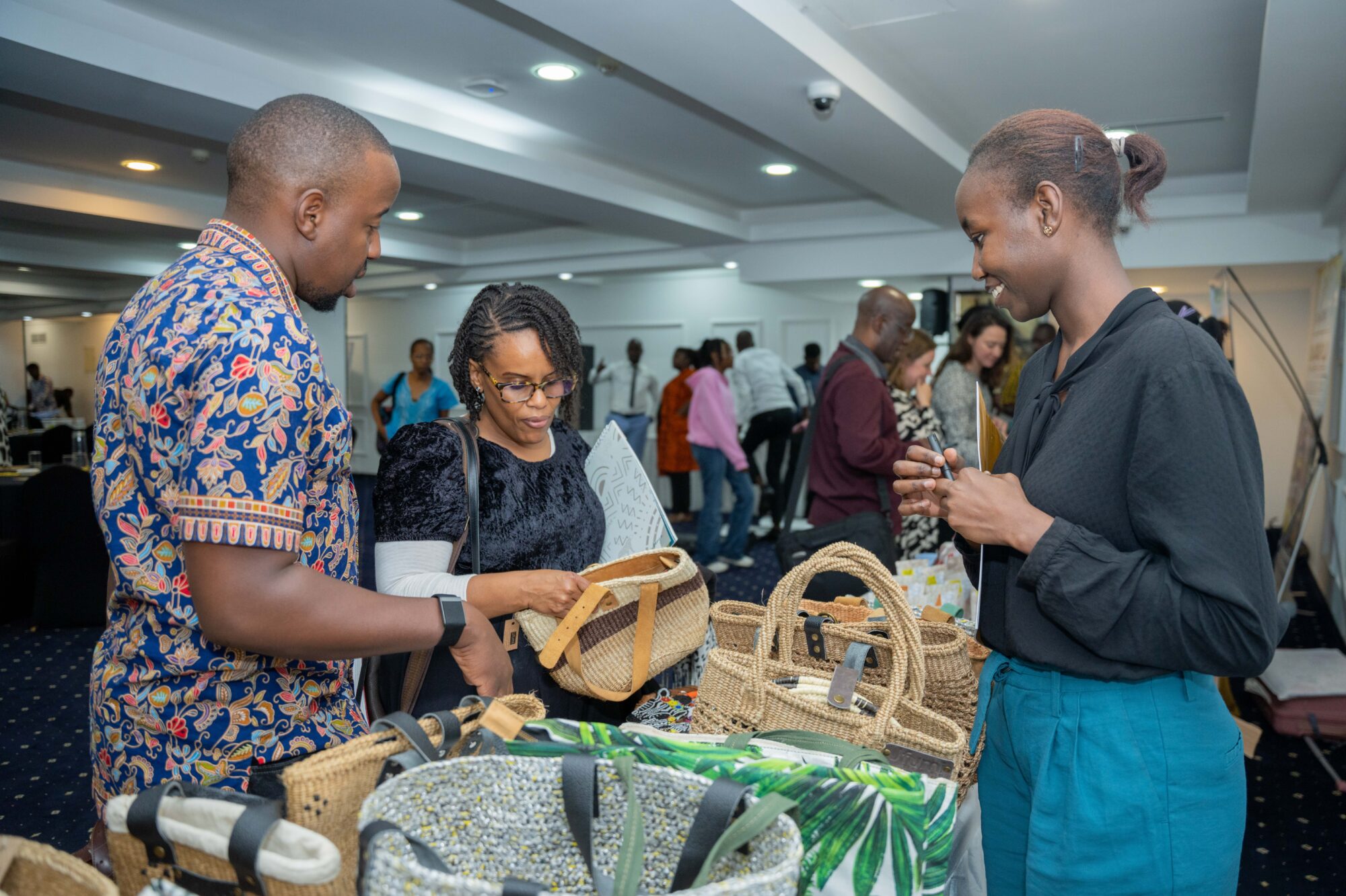 A woman stands at a display table covered in straw baskets, inside a conference-style space. She is Black with her hair in a bun and wearing a grey blouse and blue trousers, and she is smiling at two people who are standing in front of her stall looking at her products. There are more people in the background looking at other stalls.