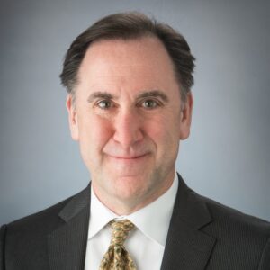 Jim Jones, a man in a black suit, white shirt, and yellow patterned tie, poses in front of a grey background. It is a professional headshot photo from his chest up. 