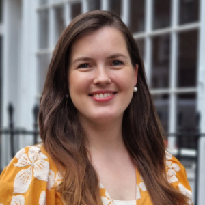 Franziska Wilk headshot. She is wearing an orange dress with white botanical patterns on it. Her hair is long and brown. The photo is framed from her shoulders up. She is outside and standing in front of a window and fence. 