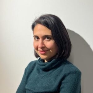 Ioanna Nitsou, Head of Principle Donors at the Cherie Blair Foundation for Women, stands in front of a white wall. She wears a blue-green turtle neck and is stood with her body angled slightly to the left. She has dark hair that falls at her jawline. She is smiling. 