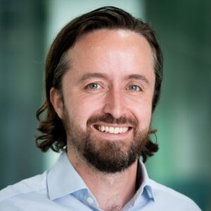 John Bruce, Head of Finance at the Cherie Blair Foundation for Women, stands in front of a green background. The image is a headshot, showing him from his shoulders up. He is wearing a blue button down shirt. He has brown, medium length hair and a beard. He is smiling. 