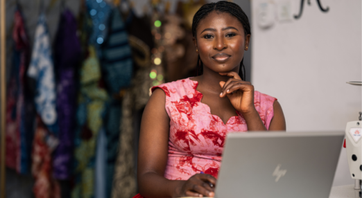 Woman entrepreneur sits at a table with a sliver laptop open on the top surface. She is wearing a pink and red dress. One hand is on the computer while the other is touching her face. In the background is a rack of vibrant and colourful clothing.
