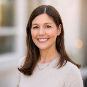 Rachel Seftel, Head of Research & Impact at the Cherie Blair Foundation for Women stands smiling in front of a white building on a city street. She has straight brown hair that falls just below her shoulder. She is wearing a tan shirt and a simple necklace. The photo is taken from a slight angle with her head turned toward the camera. It is shot from the chest up. 